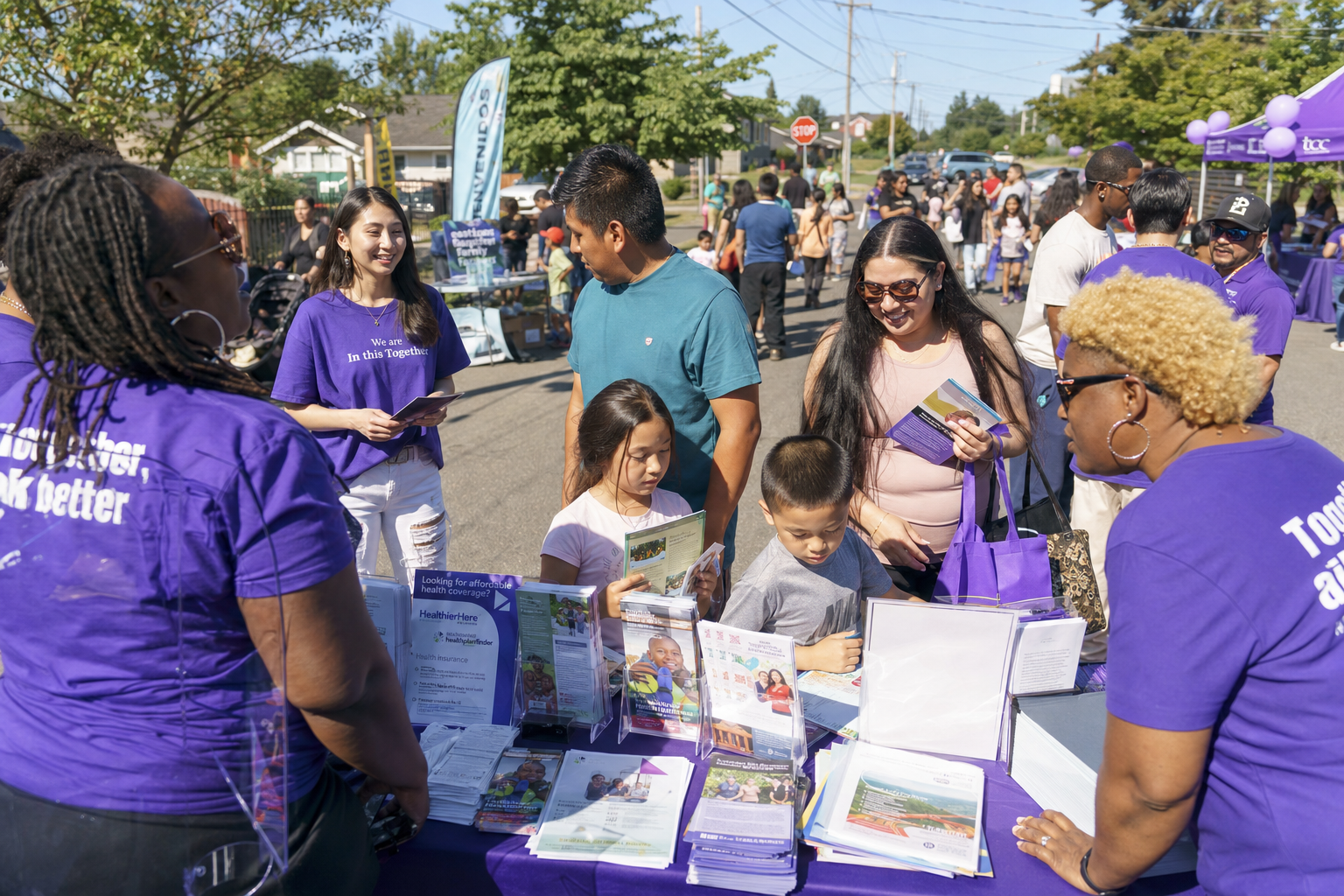 A busy community outreach event in a communities across the Greater Puget Sound region, including King, Pierce, and Snohomish Counties neighborhood, with diverse families gathering at resource tables staffed by volunteers in purple shirts